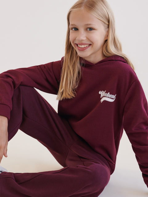 Young girl wearing a maroon hoodie with a logo, sitting on a white background