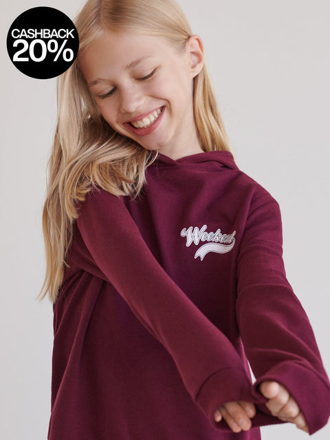 Young girl wearing a maroon hoodie with a logo on a plain background