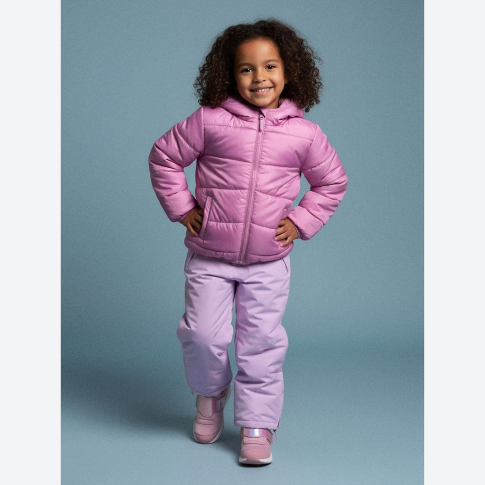 A young girl with curly dark hair stands smiling in a studio, wearing a pink hooded puffer jacket and matching light purple-pink snow pants with pink shoes, against a solid blue-gray background.