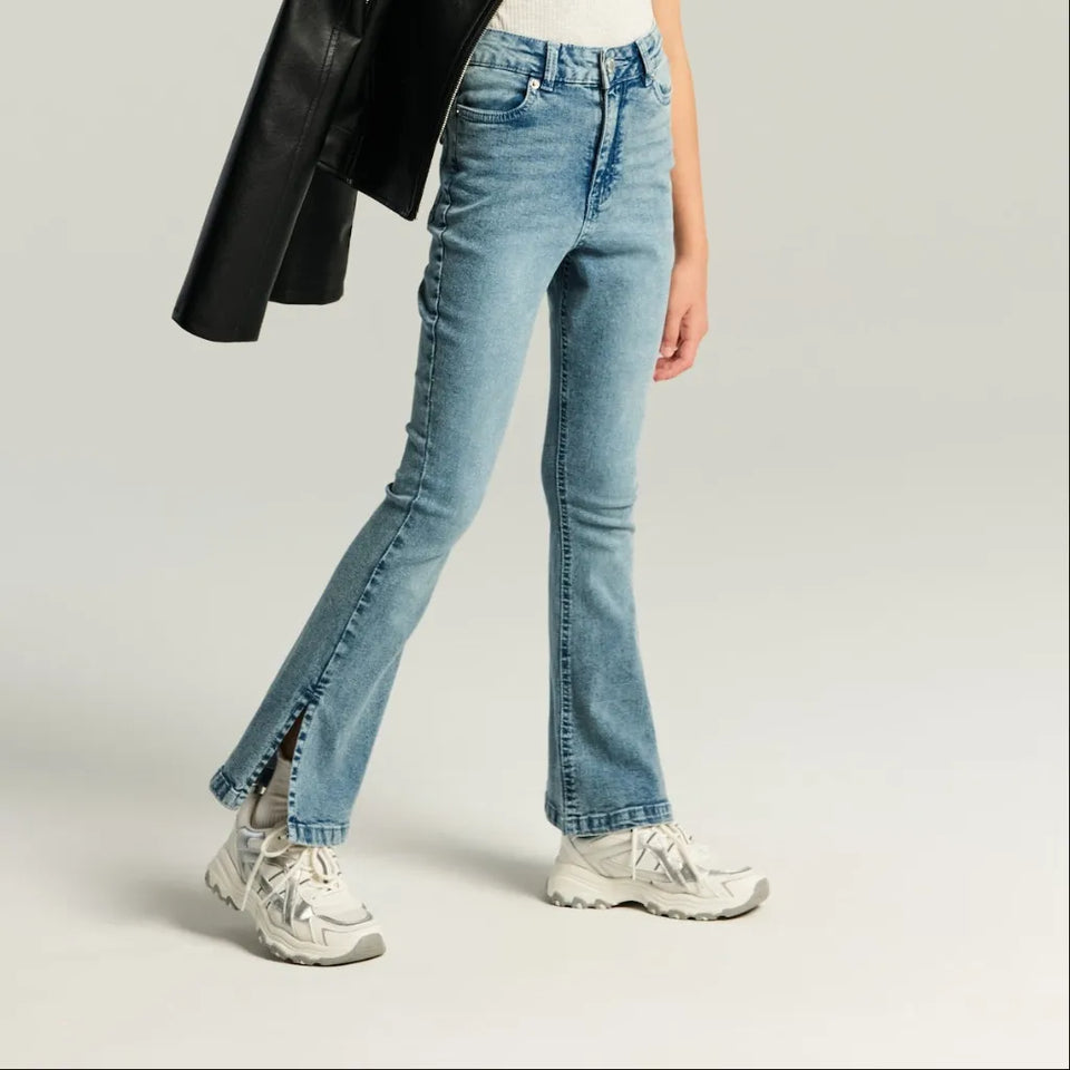 A model wearing light-wash blue flare jeans with side slits, styled with white sneakers and a black jacket, shown from the waist down on a light gray studio background.