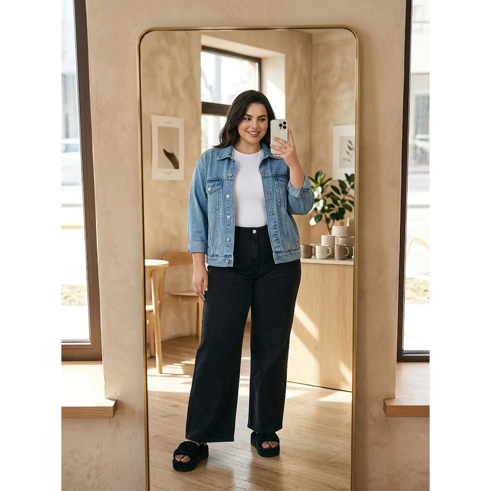 A woman takes a mirror selfie in a stylish room, wearing a light-wash blue denim jacket over a white top, black wide-leg pants, and black slide sandals.