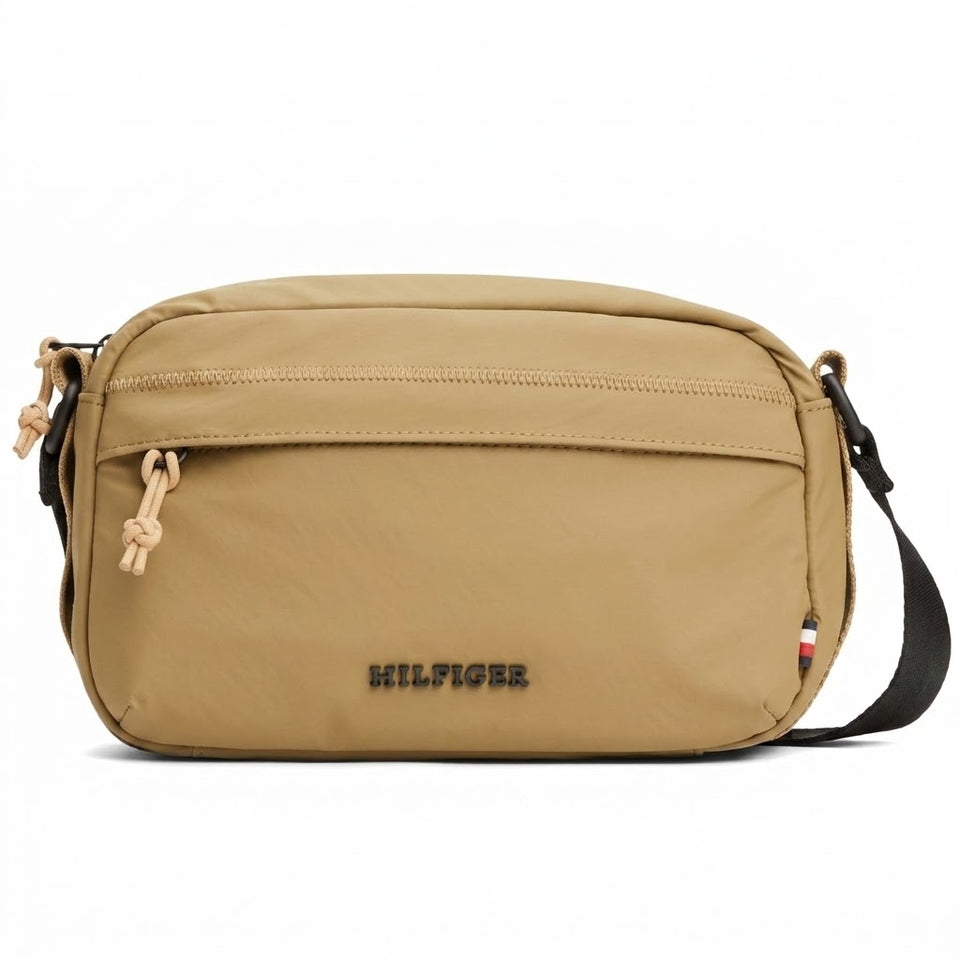 A tan nylon crossbody bag with a black adjustable strap, featuring the black embroidered 'HILFIGER' logo on the front and a small red, white, and blue flag detail on the side.