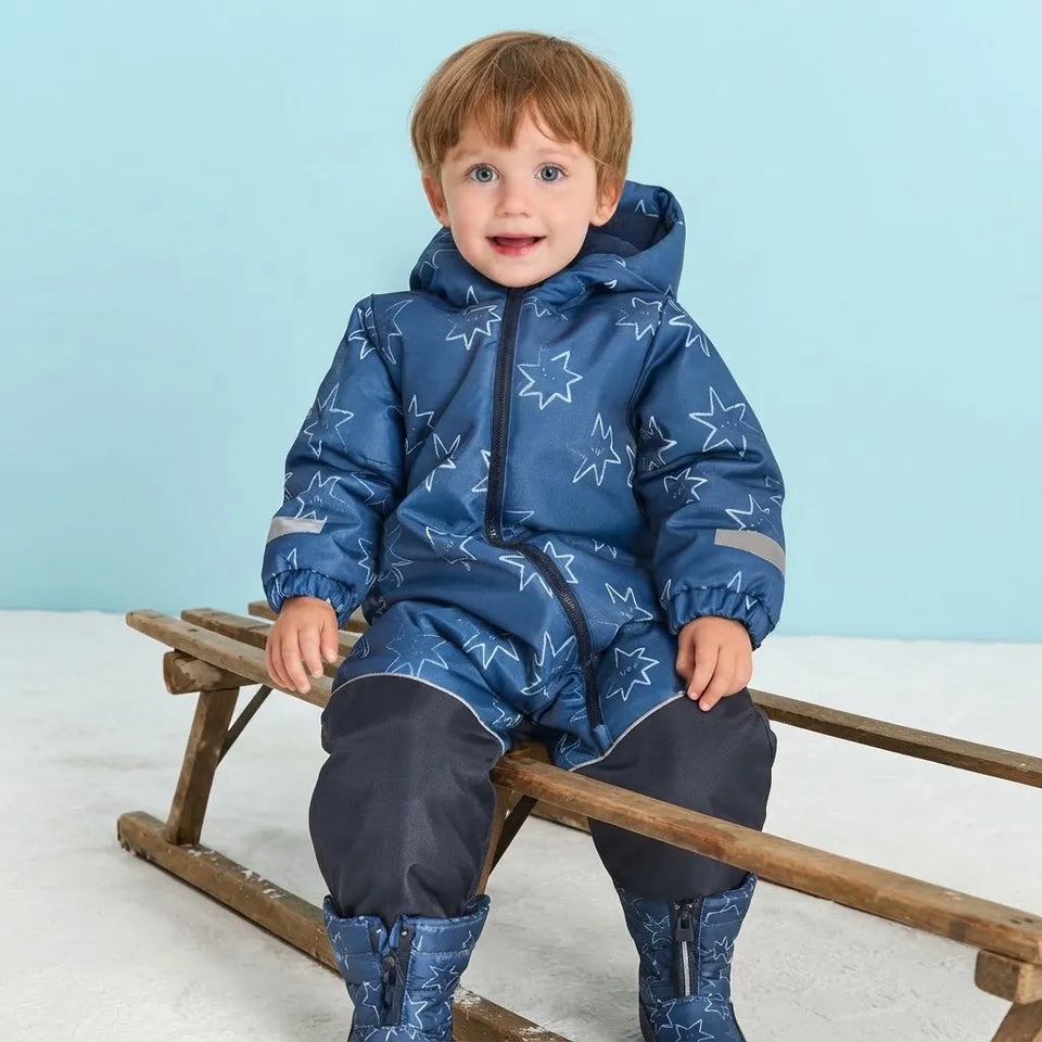 A young child wearing a blue hooded snowsuit with a white star pattern, sitting on a wooden sled against a light blue background.
