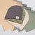 Three soft fabric beanie hats stacked on a geometric pastel background. The top hat is dark gray, the middle is olive green with a line-drawn pattern, and the bottom is light beige.