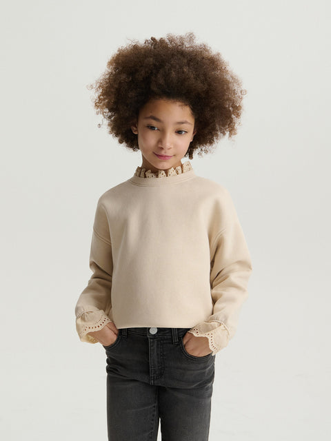 Child wearing a beige sweater with lace details on a plain background