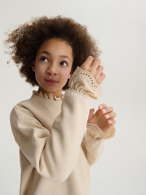 Child wearing a beige sweater with lace details on a plain background
