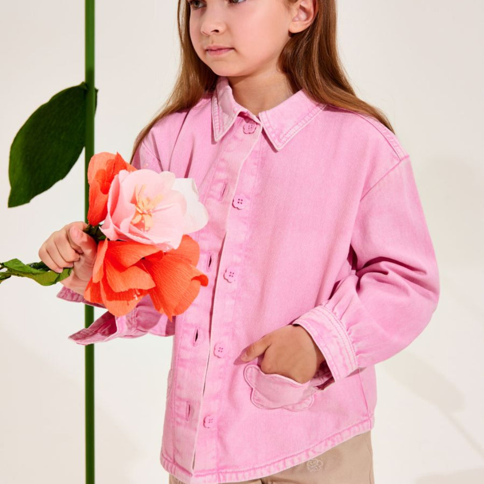 A young girl with long light brown hair wearing a pink button-up shirt and beige pants, holding a bouquet of orange and pink flowers next to a giant yellow flower prop against a plain light background.