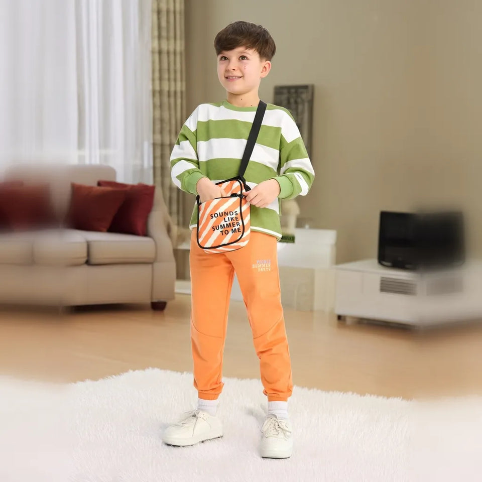 A young boy stands in a living room wearing a green and white striped long-sleeve shirt, orange sweatpants, and white sneakers, while carrying a small crossbody bag with orange and white diagonal stripes and black text.