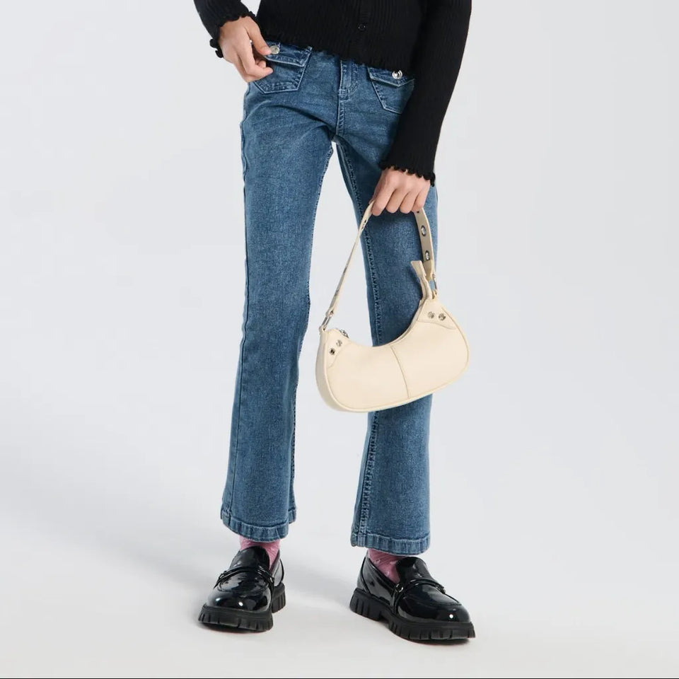 A model wearing blue flare jeans, a black sweater, and black patent leather loafers, holding a cream-colored crescent-shaped shoulder bag with a gold-tone strap.