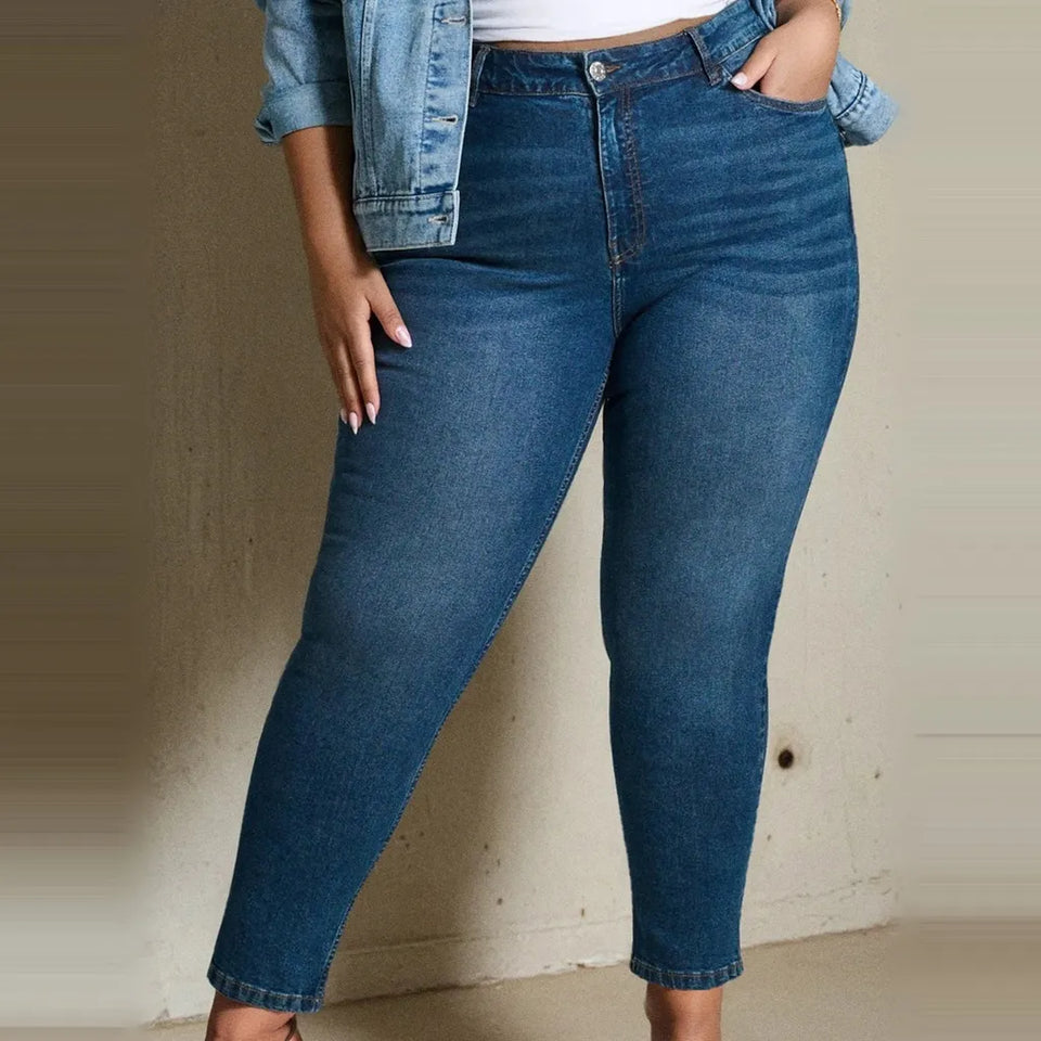 A person wearing high-waisted, ankle-length blue denim jeans with a fitted silhouette, styled with a light blue denim jacket and white top against a neutral indoor wall.