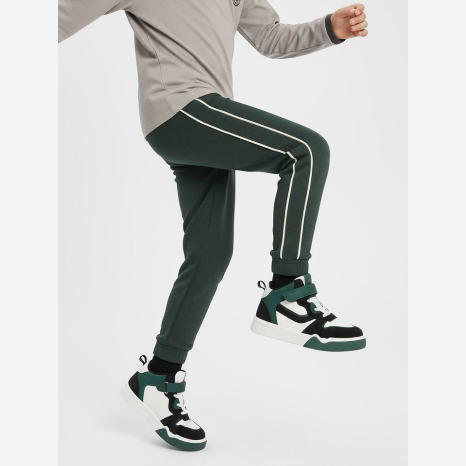A person wearing dark green jogger-style pants with white side stripes, paired with a light gray long-sleeve top and black-and-white sneakers, captured mid-motion against a plain light gray studio background.