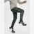 A person wearing dark green jogger-style pants with white side stripes, paired with a light gray long-sleeve top and black-and-white sneakers, captured mid-motion against a plain light gray studio background.