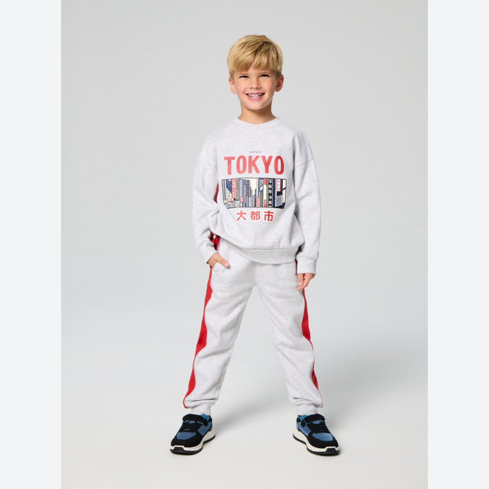 A young boy stands against a light gray studio background wearing a light gray sweatshirt and matching sweatpants with red side stripes and a red 'TOKYO' graphic on the chest, paired with dark blue sneakers.