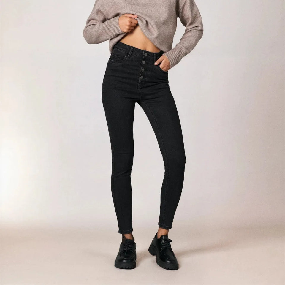 A model wearing high-waisted black skinny jeans with a button-fly front, styled with a cropped gray knit sweater and black leather shoes against a neutral studio backdrop.