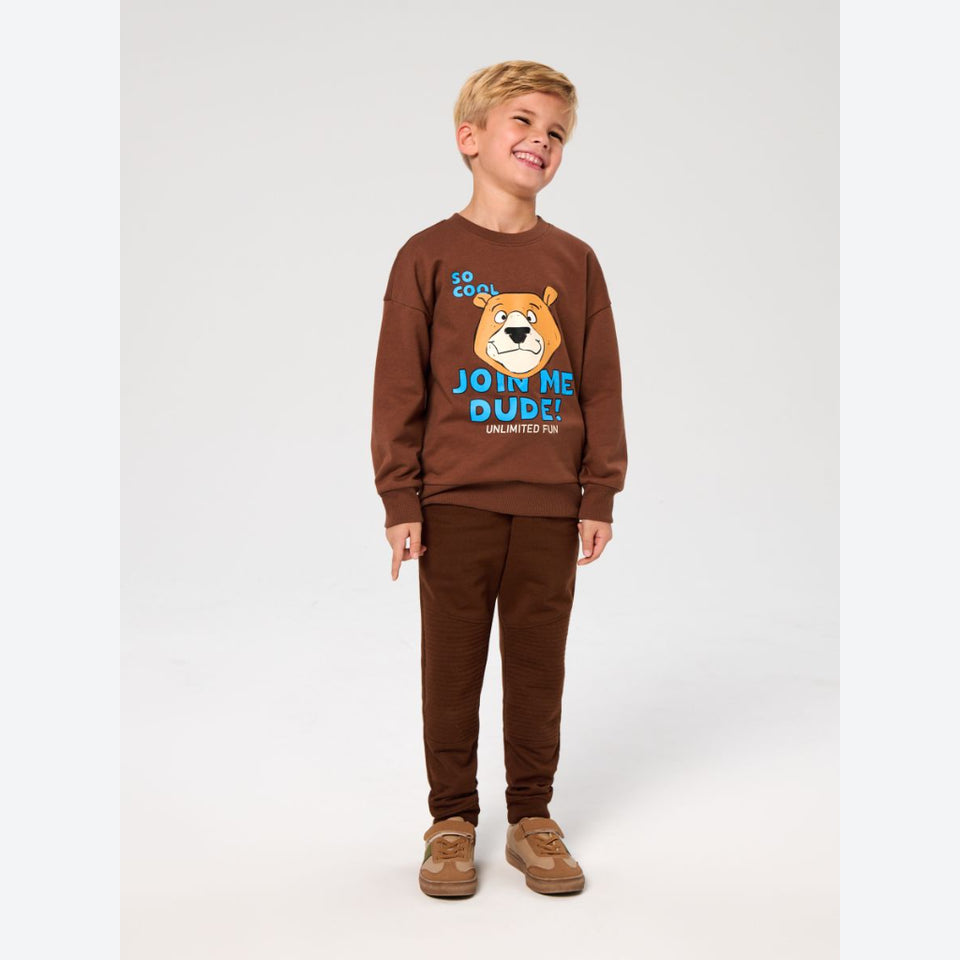 A young boy stands against a light gray studio background wearing a matching brown sweatshirt and sweatpants set. The sweatshirt features a colorful graphic of a cartoon bear face with the text 'SO COOL JOIN ME DUDE! UNLIMITED FUN'.