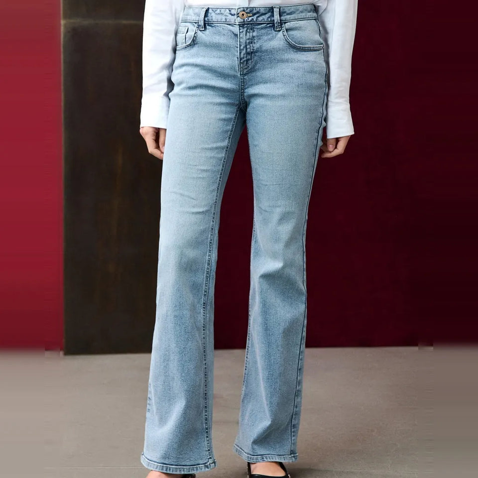A person wearing light-wash blue flare jeans with a white long-sleeve top, shown from the waist down against a dark red and brown studio backdrop.