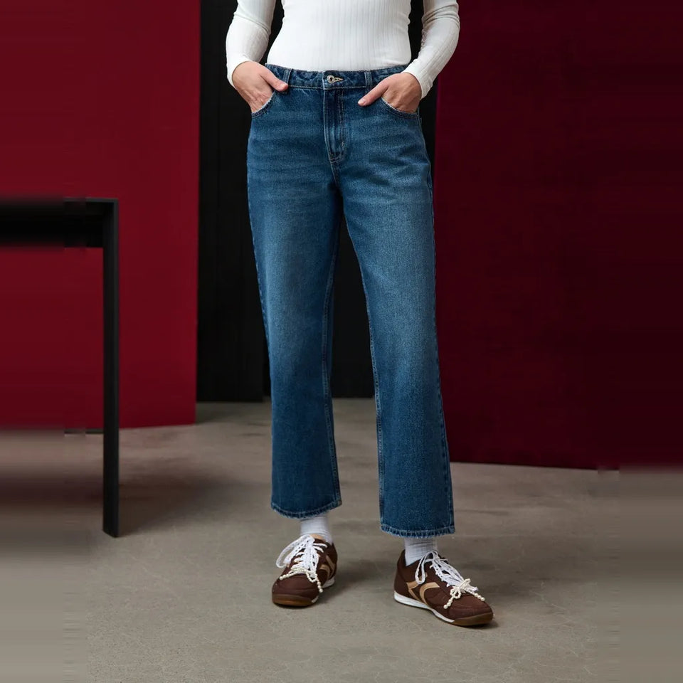 A person wearing medium-wash blue straight-leg jeans, a white ribbed long-sleeve top, white socks, and brown sneakers, standing in a studio with a dark red wall and gray floor.