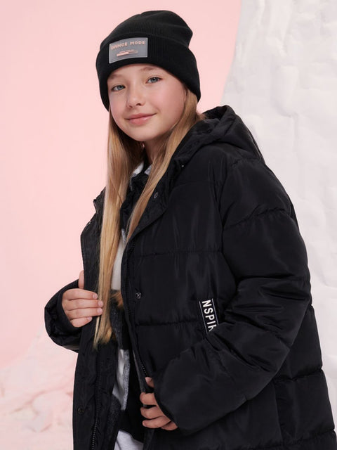 Young girl wearing a black jacket and beanie with visible branding against a pink background
