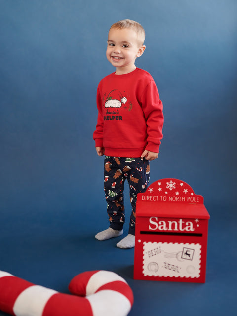 Child wearing a red 'Santa Helper' sweater and patterned pants, standing next to a red Santa mailbox on a blue background.