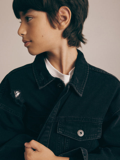 Young person wearing a dark denim jacket over a white shirt against a neutral background