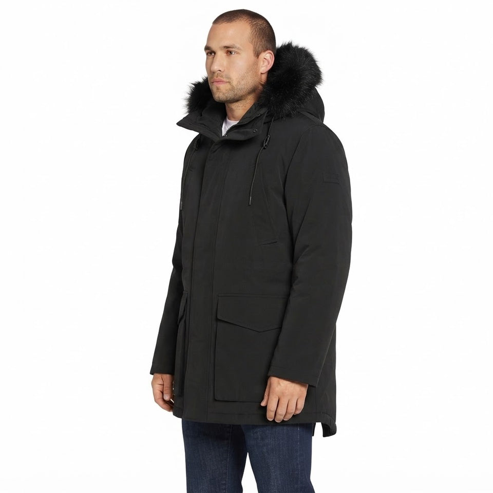 A man wearing a black hooded parka with a dark fur-trimmed hood, shown in profile against a plain white studio background.