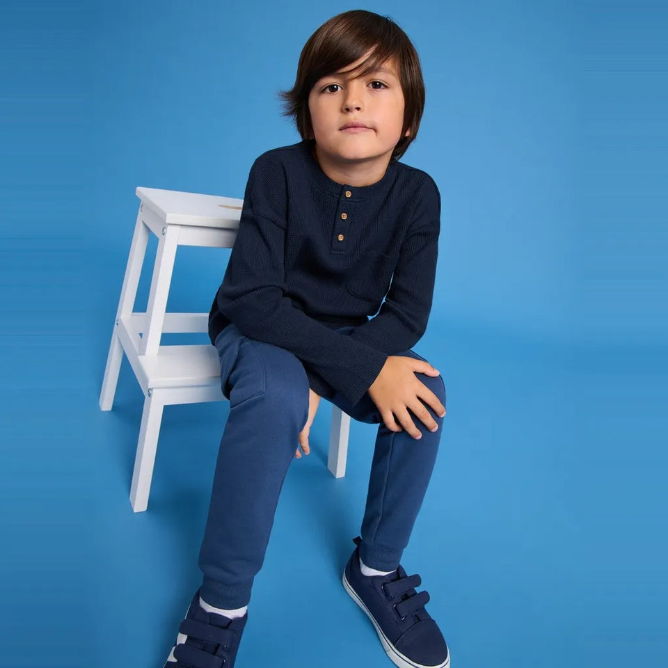 A young boy wearing a navy blue long-sleeve henley shirt, navy pants, and navy sneakers sits on a small white wooden stool against a solid blue studio background.