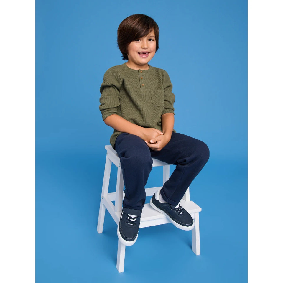 A young boy with brown hair sits smiling on a small white wooden step stool against a solid blue studio background. He is wearing an olive green long-sleeve henley shirt, dark navy pants, and navy sneakers with white laces.