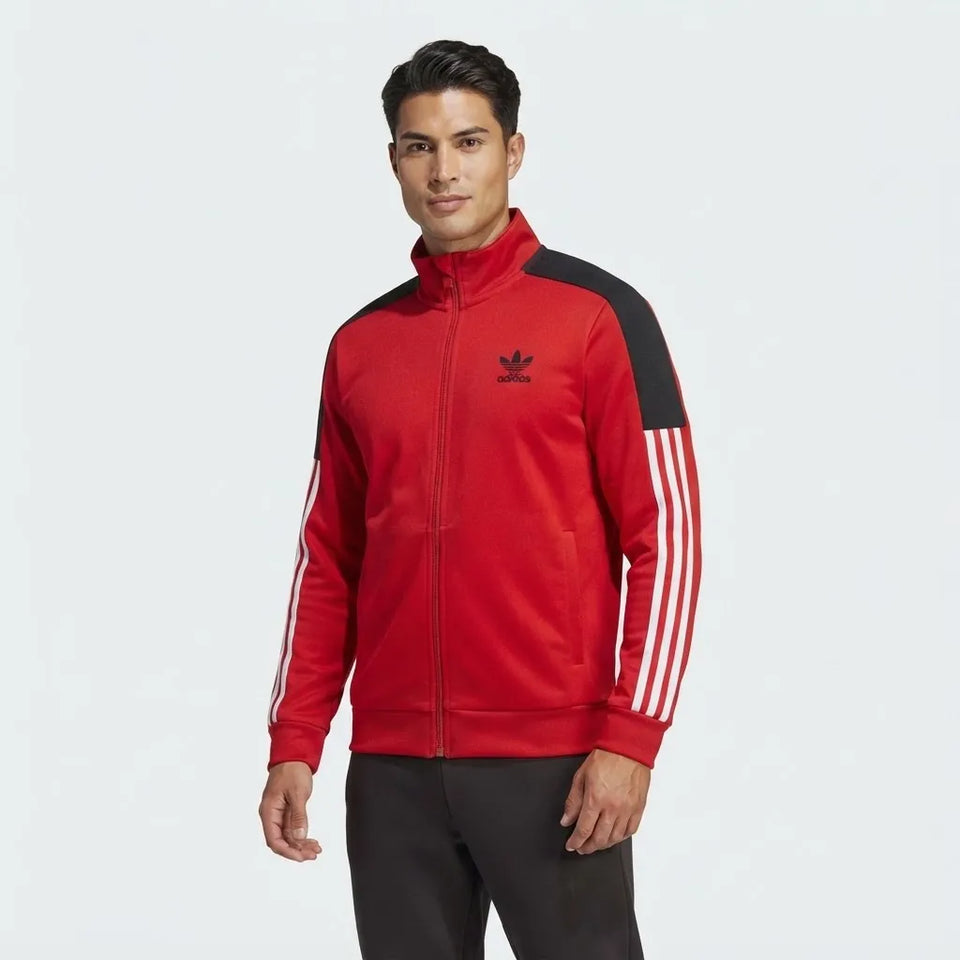 A man wearing a red Adidas track jacket with black shoulder panels, white three-stripes on the sleeves, and a black trefoil logo on the chest, set against a plain light gray background.