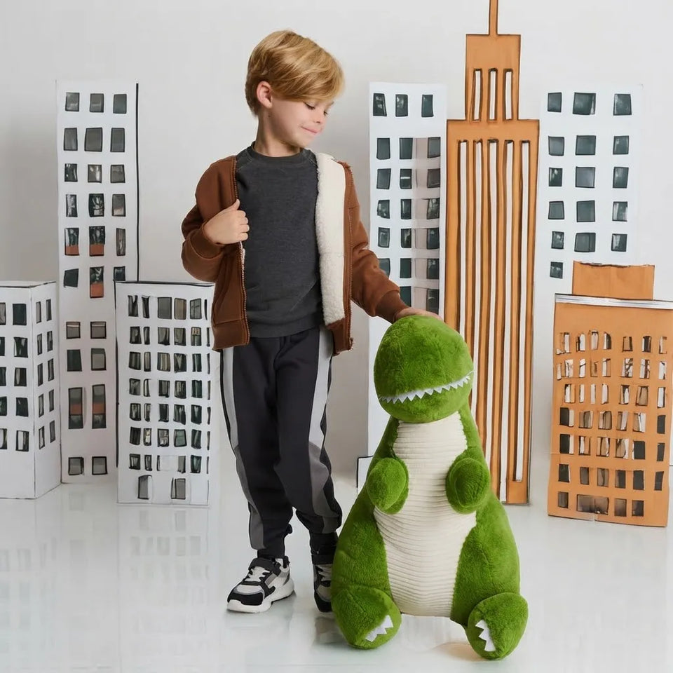 A young boy stands next to a large green plush dinosaur toy in a room decorated with cardboard city buildings.