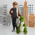 A young boy stands next to a large green plush dinosaur toy in a room decorated with cardboard city buildings.
