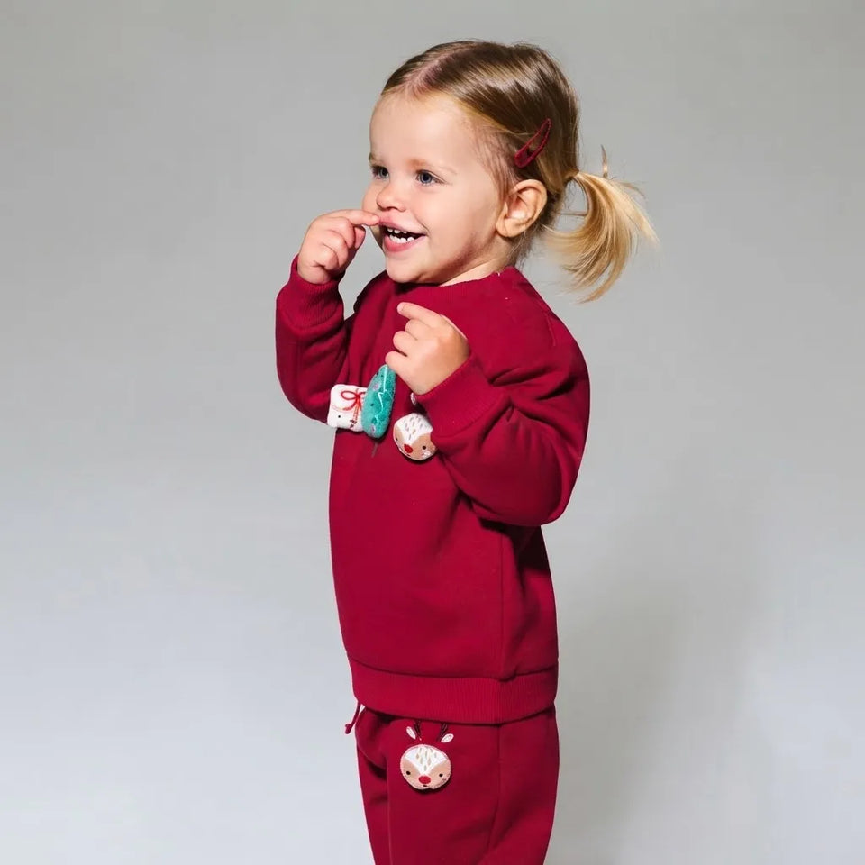 A young girl with blonde hair in a ponytail smiles while wearing a matching burgundy red two-piece pajama set with cute animal appliques, photographed against a neutral gray studio background.