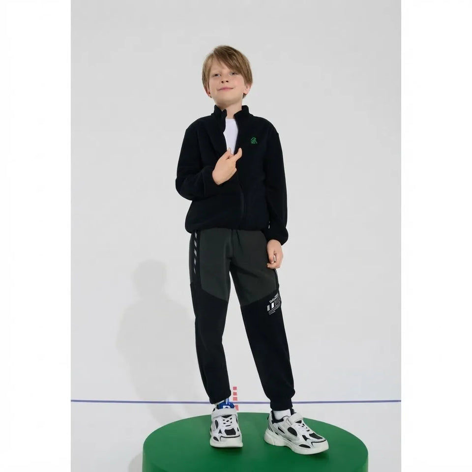 A child model wearing a black zip-up fleece jacket, black cargo pants, and white sneakers, standing on a green cylindrical pedestal against a light gray studio background.