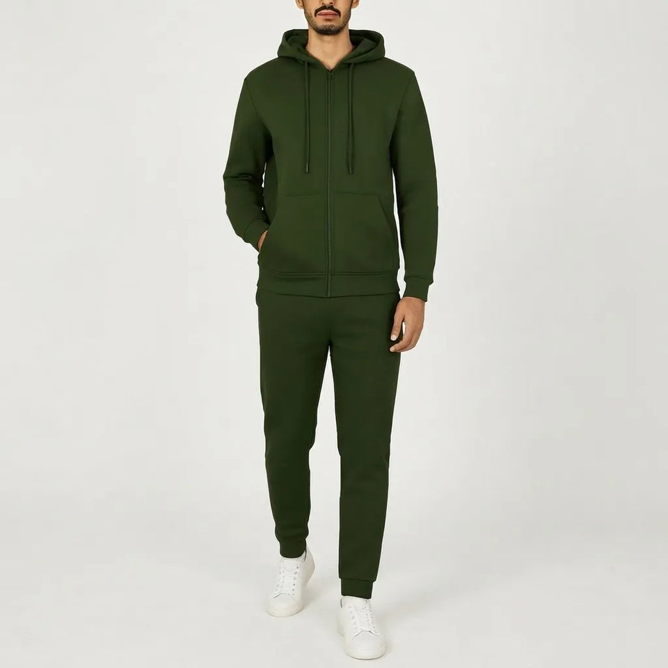 A man wearing a matching dark green zip-up hoodie and sweatpants set, paired with white sneakers, standing against a plain light gray studio background.