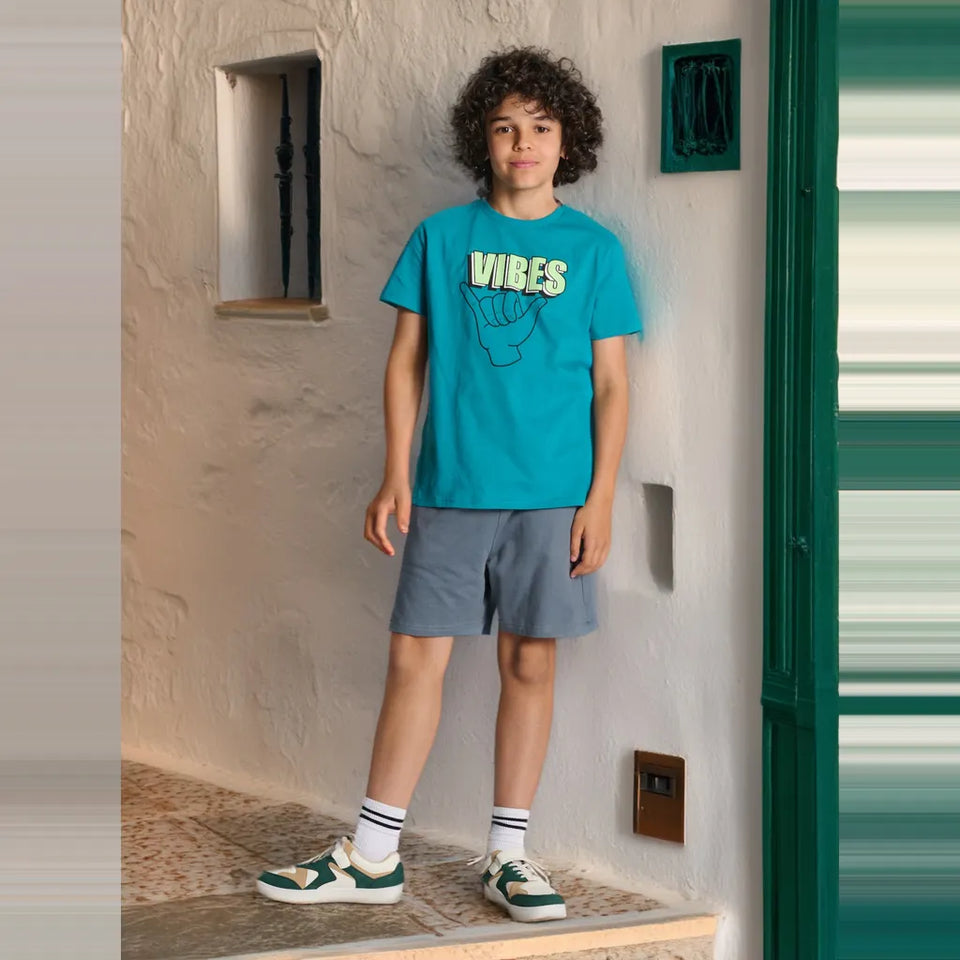 A child model wearing a bright turquoise graphic t-shirt with 'VIBES' and a hand graphic, paired with slate blue shorts, white socks, and green and white sneakers, standing in a doorway with textured white walls.