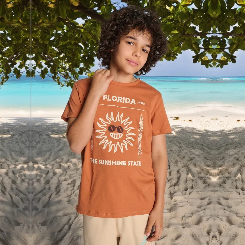 A child wearing a terracotta orange graphic t-shirt with a white sun design and 'FLORIDA' text, standing on a sandy beach with turquoise water and a tree canopy in the background.
