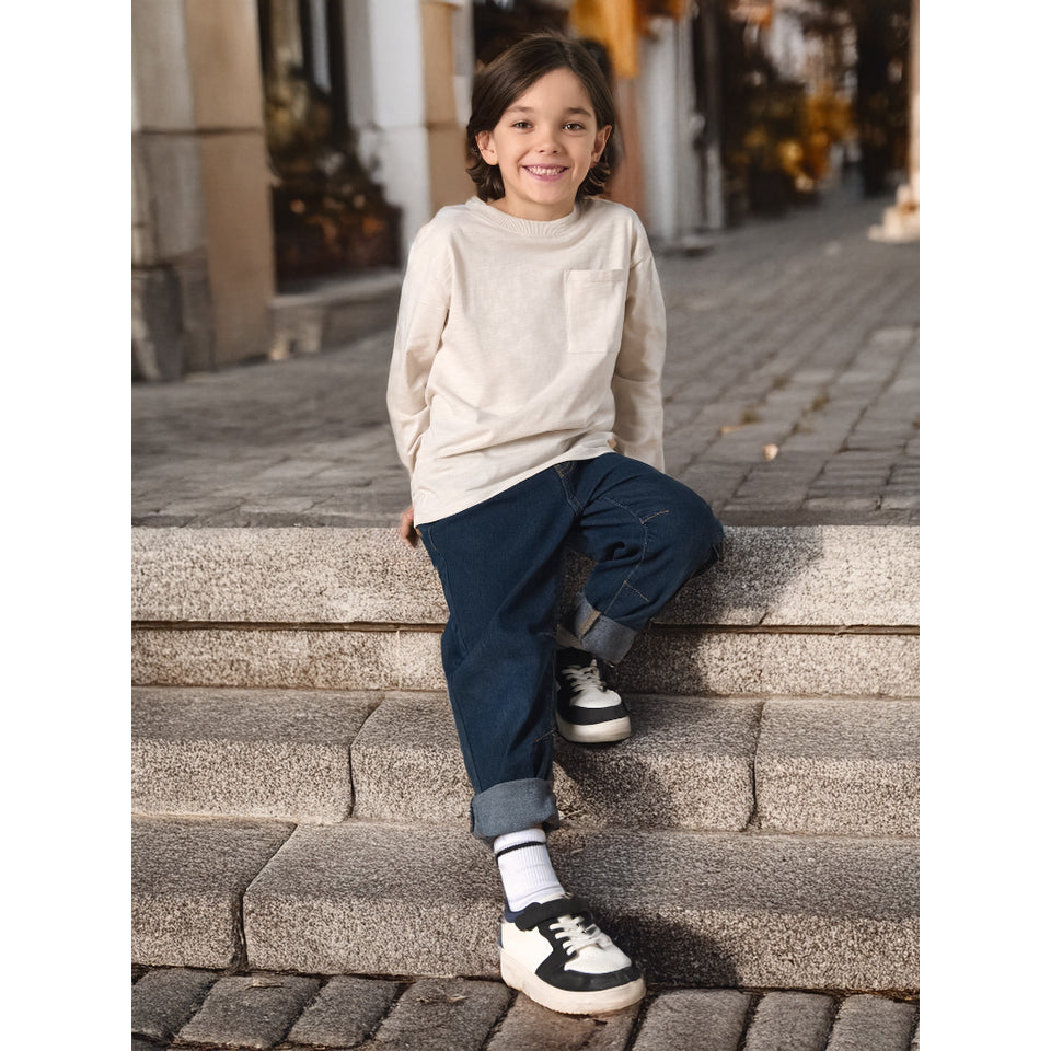 A young child sitting on stone steps outdoors, wearing a cream long-sleeve pocket top, dark blue jeans, white socks, and black-and-white sneakers.