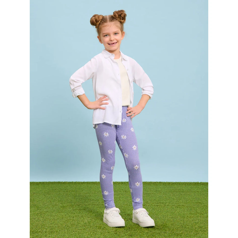 A young girl with her hair in two buns stands on a green grass-like surface against a light blue background, wearing a white button-up shirt over a cream top, purple-and-white gingham leggings with white floral prints, and white sneakers.
