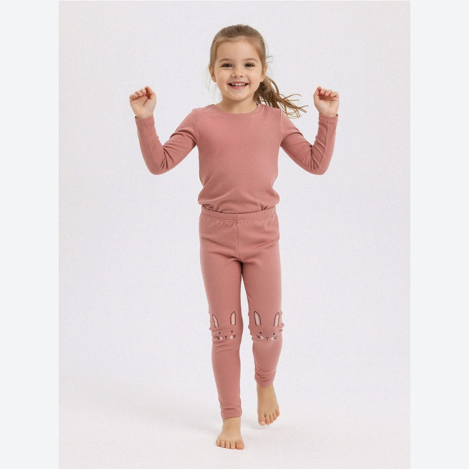 A young girl smiling and jumping barefoot in a matching dusty rose long-sleeve top and jogger pants with bunny ear details on the legs, set against a plain light gray studio background.