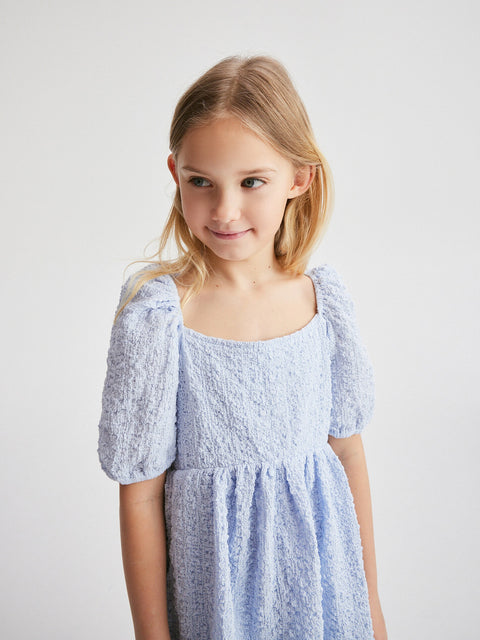 Young girl wearing a light blue dress with puff sleeves against a plain background