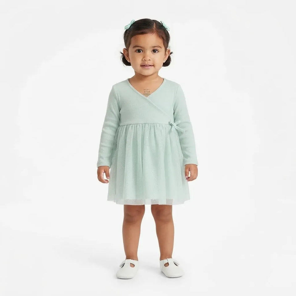 A toddler girl wearing a mint green long-sleeve tulle dress with a crossover V-neck and a small bow detail at the waist, standing against a plain white studio background.