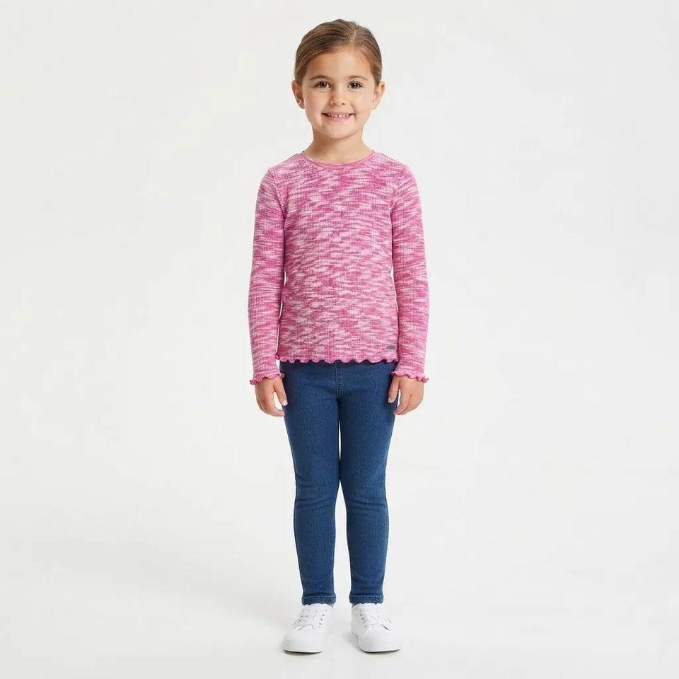 A young girl stands against a plain white studio background wearing a pink and white marled knit long-sleeve top with a scalloped hem, blue skinny jeans, and white sneakers.
