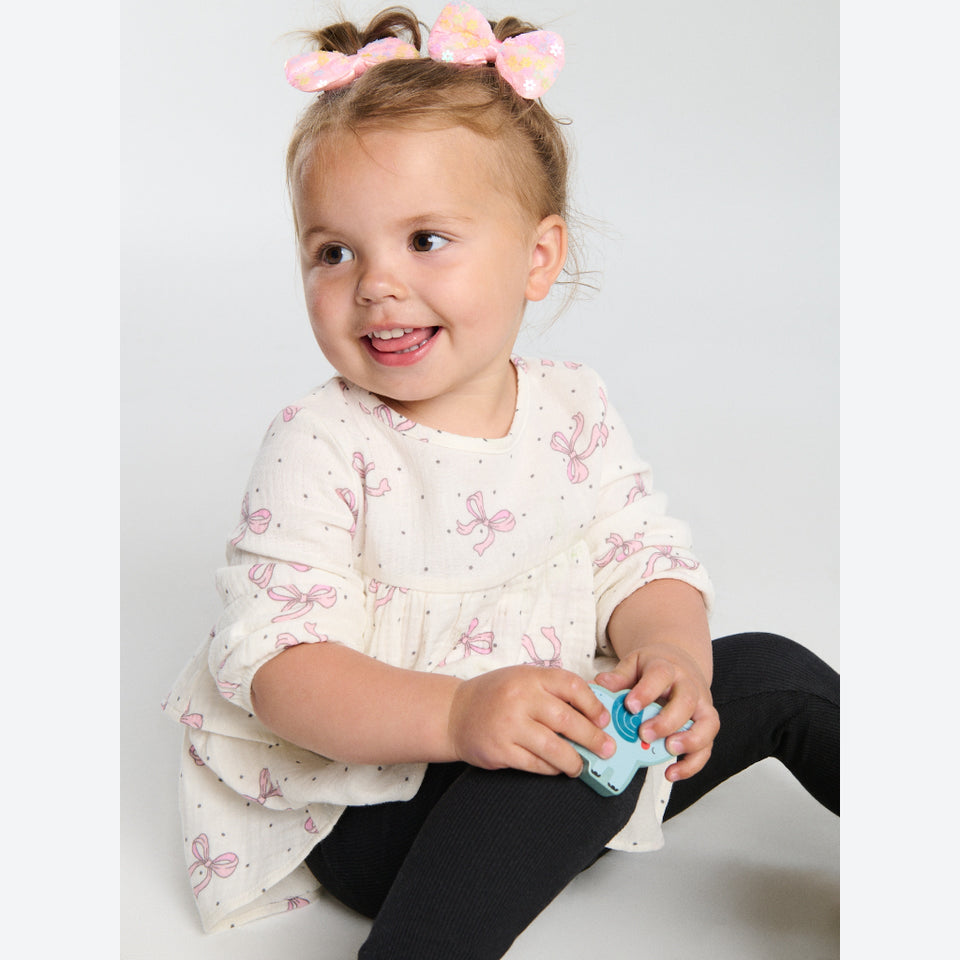 A toddler girl sitting on a light gray studio floor, wearing a cream long-sleeve top with a pink bow and butterfly print, black leggings, and a pink bow in her hair, holding a small blue toy.