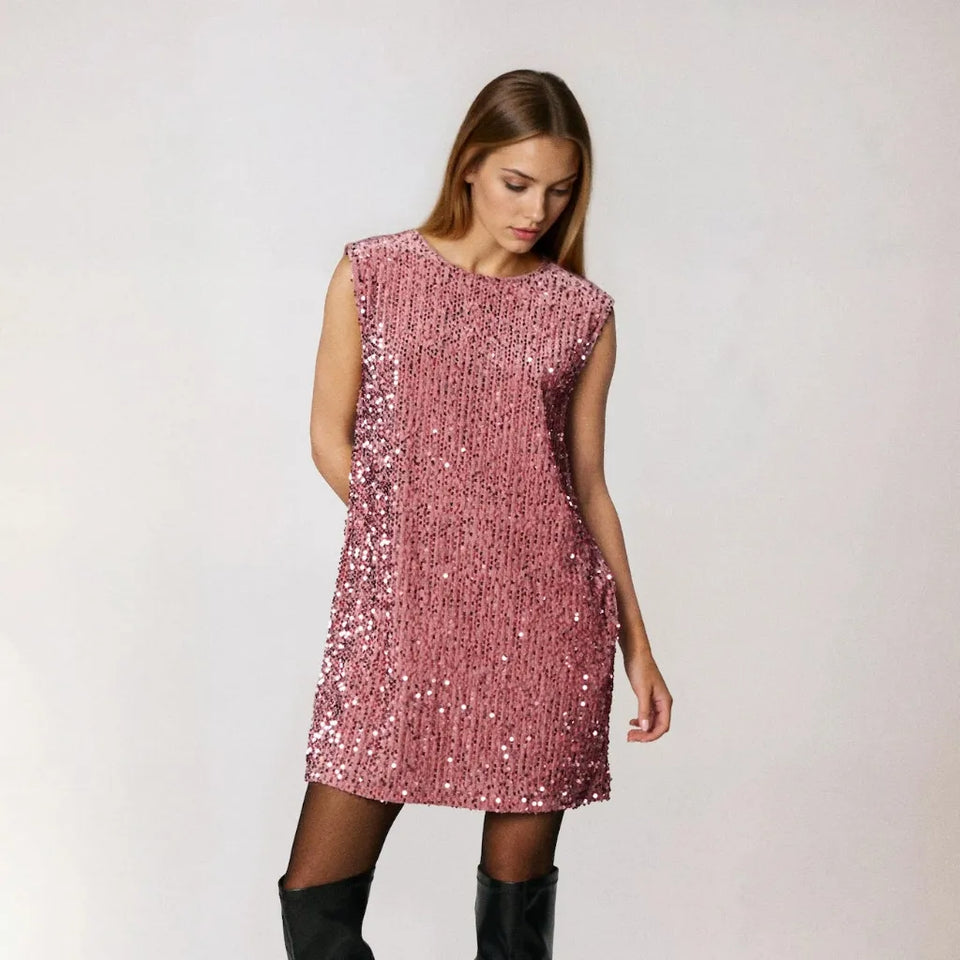 A woman modeling a sleeveless, pink sequined mini dress paired with black thigh-high stockings and black knee-high platform boots, photographed against a neutral light gray studio background.