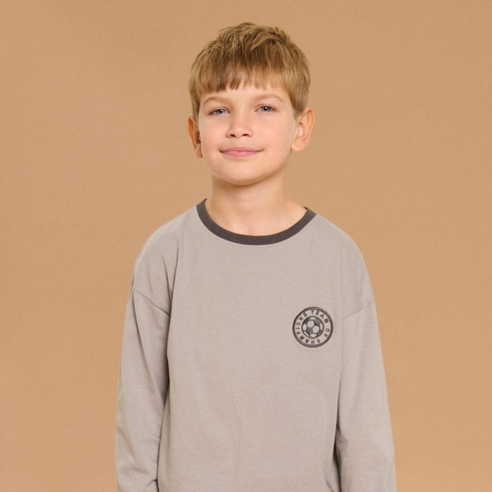 A young boy wearing a light gray long-sleeve t-shirt with dark gray ribbed collar, cuffs, and a circular chest graphic, paired with light gray sweatpants, set against a solid warm beige background.