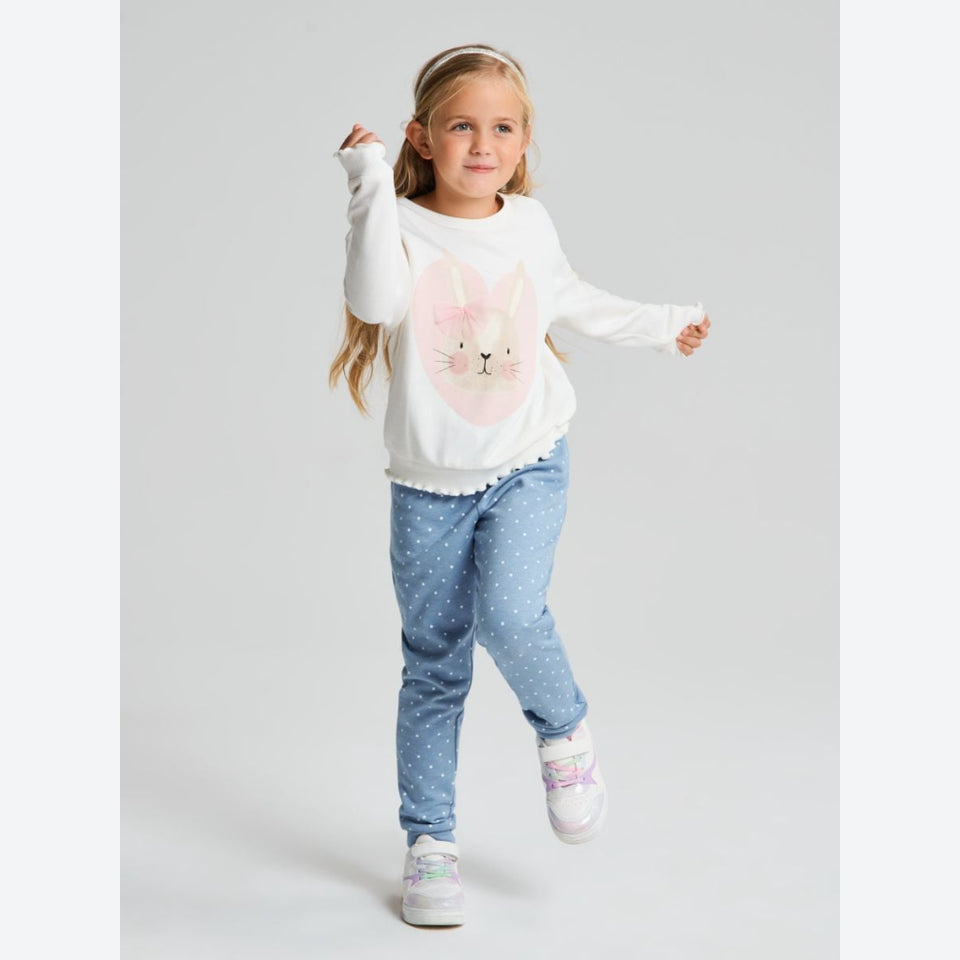 A young girl wearing a white long-sleeve sweatshirt with a pink bunny face graphic, light blue patterned pants, and white sneakers, posing against a light gray studio background.