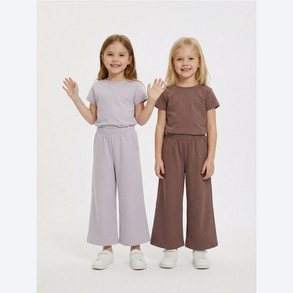 Two young girls modeling matching two-piece lounge sets. The girl on the left wears a light lavender short-sleeve top and wide-leg pants, while the girl on the right wears a medium brown short-sleeve top and matching wide-leg pants.