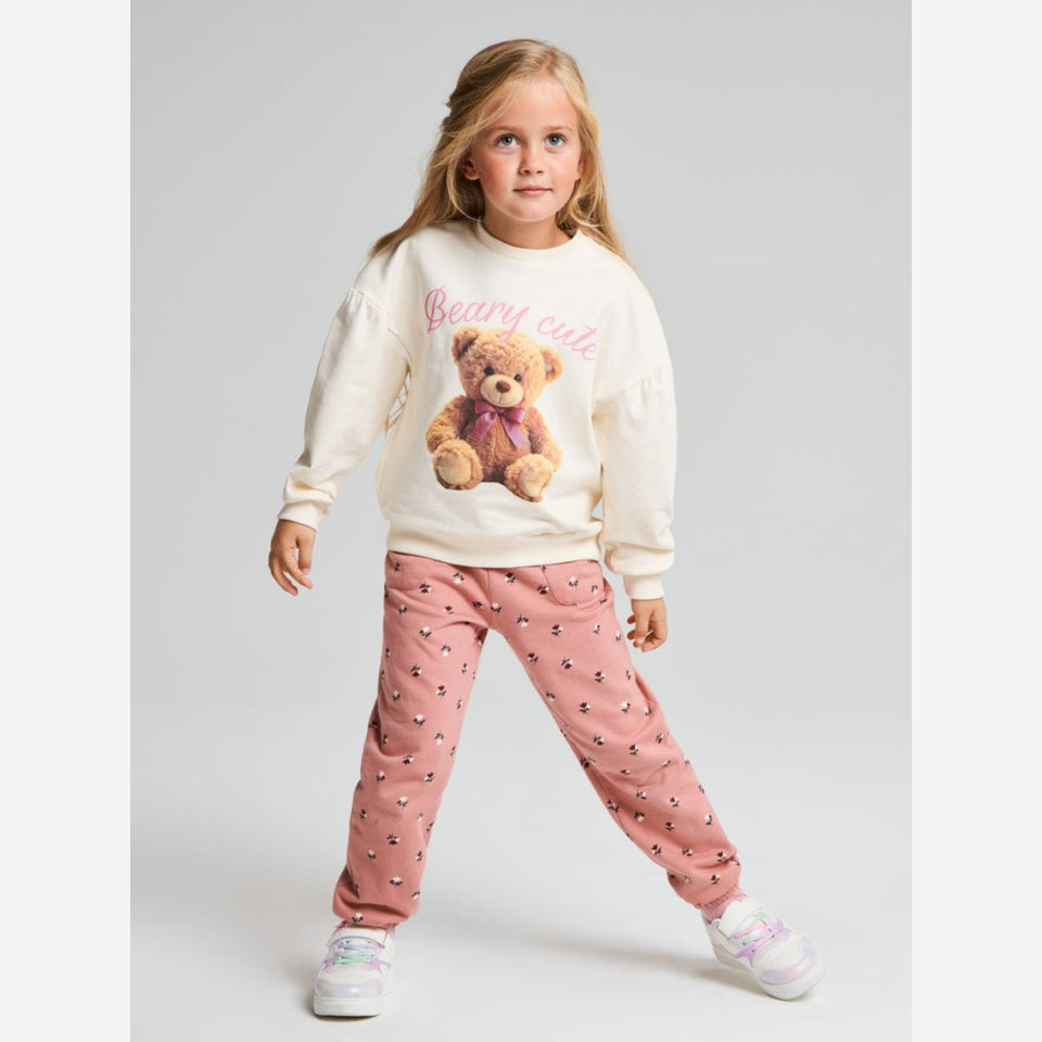 A young girl stands in a studio wearing a cream sweatshirt with a teddy bear graphic and pink pants with a small pattern, paired with white sneakers.