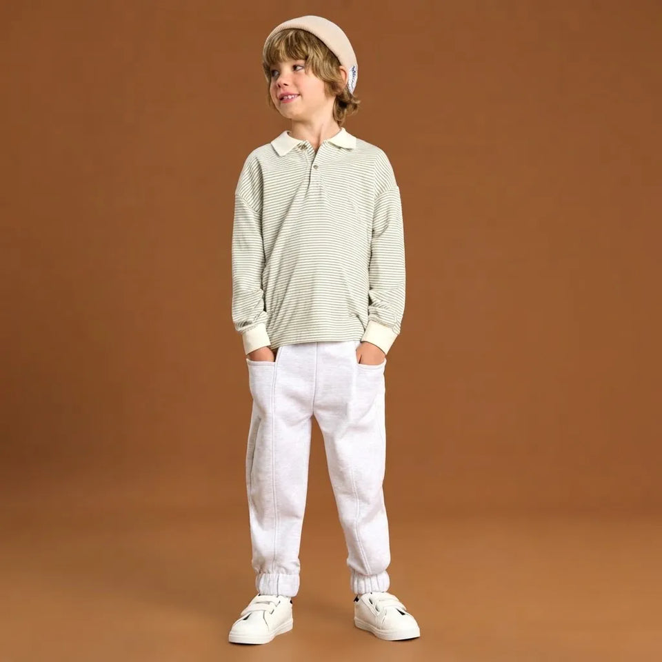 A young boy modeling a light green and white striped long-sleeve polo shirt, white pants, and white sneakers against a solid brown studio background.