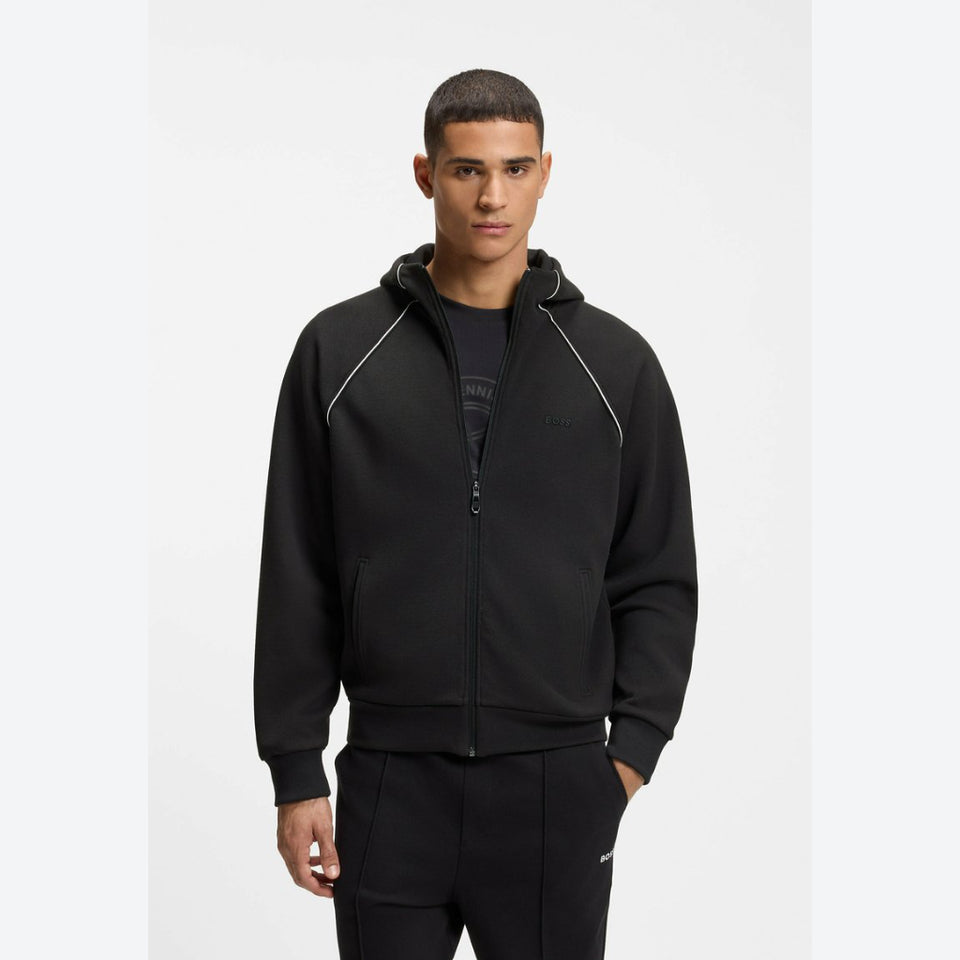 A man wearing a black zip-up hoodie with white contrast piping and a black t-shirt underneath, standing against a plain light gray studio background.