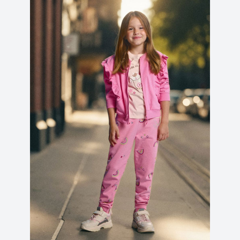 A young girl stands on a city street wearing a bright pink matching set consisting of a ruffled zip-up jacket and printed jogger pants, over a light pink graphic T-shirt and white sneakers.